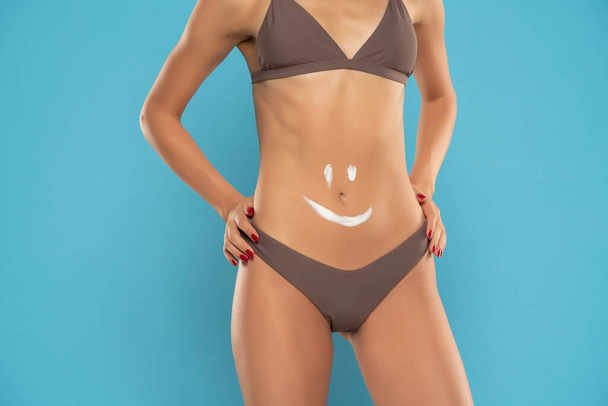 Gros plan du ventre de la femme avec le sourire dessiné avec de la lotion sur un fond bleu studio - Photo, image