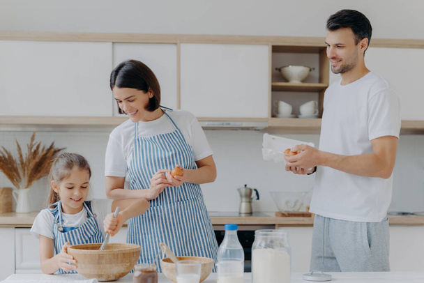 幸せな両親は娘と一緒に調理し、現代の家庭の台所でポーズをとって生地に卵を与え、小さな子供はボウルに具材を泡立て器は幸せな表情をしています。食べ物と家族の概念。朝食の調理 - 写真・画像