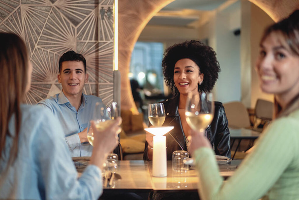 Grupa czterech przyjaciół siedzi w restauracji i szczęśliwie rozmawia trzymając szklankę białego wina. Skupiamy się na młodej Afroamerykance z kręconymi włosami, która się uśmiecha. - Zdjęcie, obraz