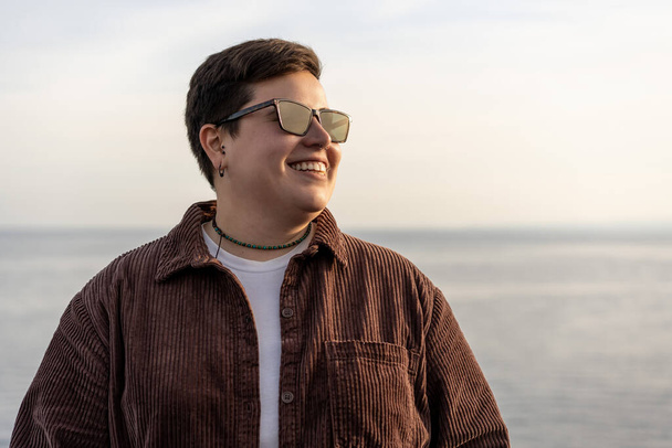 confident and joyful non-binary person with short curly hair, sporting a vintage velvet jacket and sunglasses, smiling on a scenic boardwalk during a sunny day. This portrait captures the subject from the waist up - Photo, Image