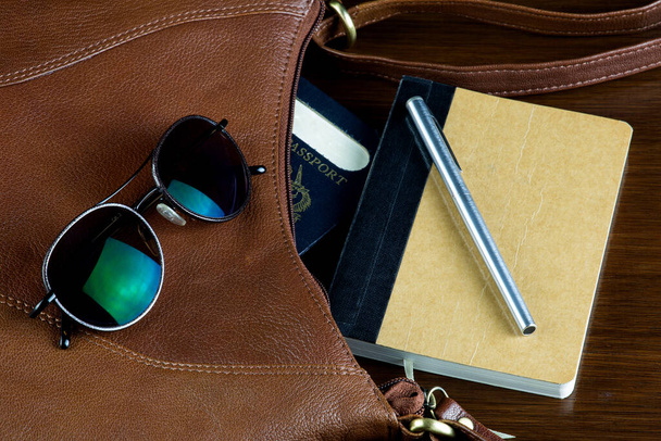 Sonnenbrille und Ledertasche mit Füllfederhalter und Notizbuch auf einer lackierten Holztischplatte - Foto, Bild