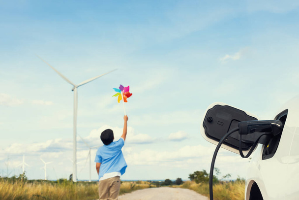 Progressive young asian boy playing with wind pinwheel toy in the wind turbine farm, green field over the hill. Green energy from renewable electric wind generator. Windmill in the countryside concept - Photo, image