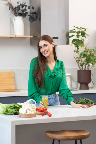 A young attractive woman is standing in the kitchen at the table with fresh vegetables and a green smoothie. Healthy eating. - Φωτογραφία, εικόνα