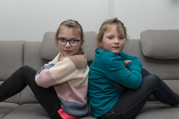 two young girls sit back to back on the sofa and look into the camera. Close up. - Foto, Imagen