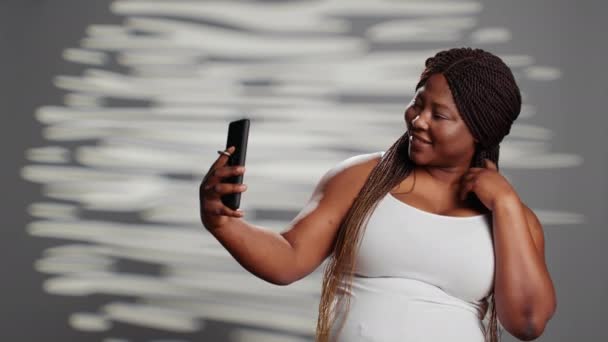 Afroamerykanka czuje się pewnie robiąc zdjęcia, za pomocą smartfona, aby pokazać swoją miłość i pozytywność ciała. Kobieta model bawiący się w studio, aby promować pewność siebie, promienną skórę. - Materiał filmowy, wideo