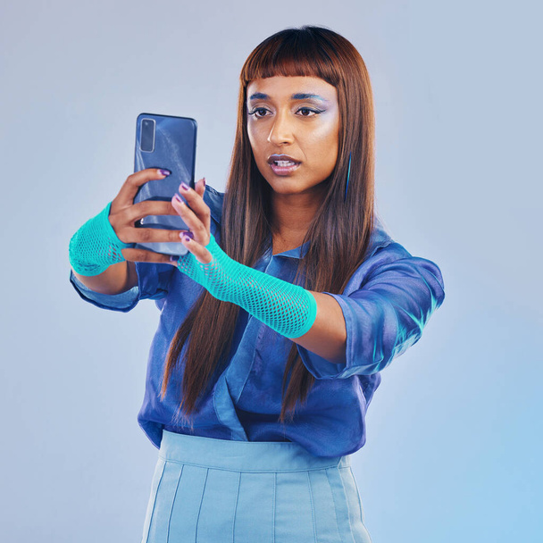 Fashion, selfie and woman with smartphone and cyberpunk neon clothing isolated on blue background. Social media, future and trendy gen z influencer from India with phone in studio for profile picture. - Foto, Imagen