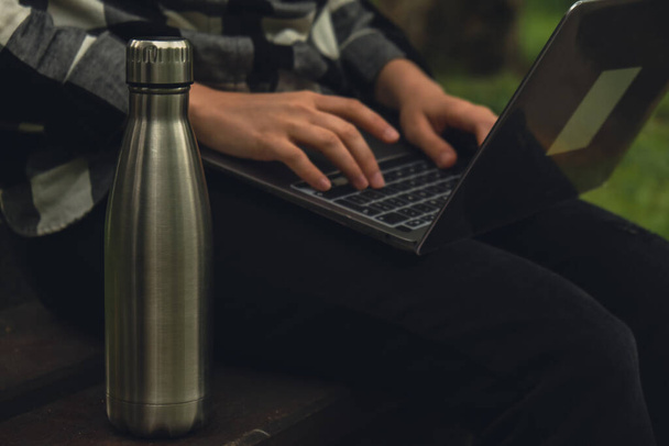 Water bottle. Reusable steel thermo water bottle on wooden bench. Sustainable lifestyle. Plastic free zero waste free living. Go green Environment protection. Woman student work study with laptop in park  - Foto, Bild