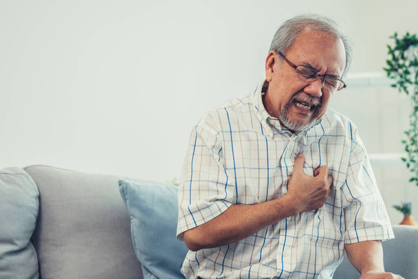 An agonizing senior man suffering from chest pain or heart attack alone in his living room. Serious health problem and feeling unwell concept for seniors. - Fotó, kép