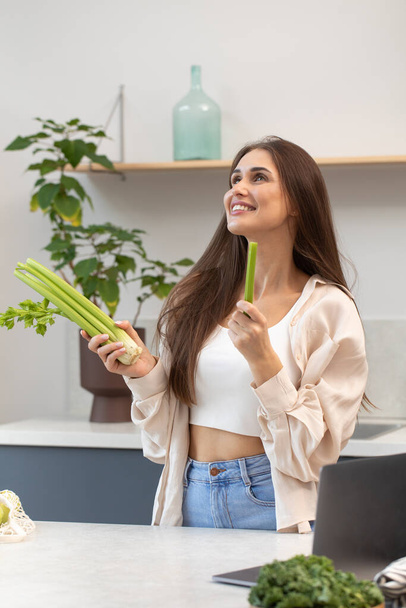 A happy woman is standing in the kitchen with fresh celery in her hands. Healthy eating, vegetarianism. - Fotó, kép
