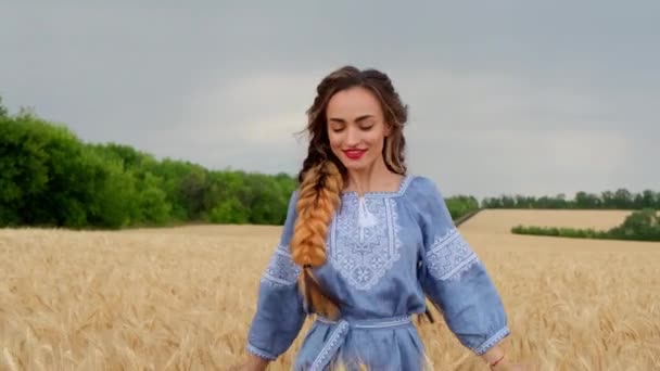 A beautiful smiling young woman in traditional Ukrainian clothes runs through the golden wheat field, spins around, enjoys outdoor recreation, freedom and carefree mood in the summer nature at sunset. - Video