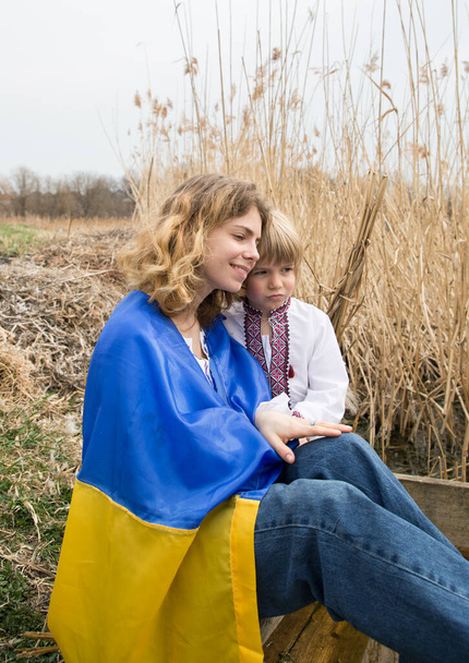 Ukrayna bayrağına sarılmış küçük bir çocuk ve bir kadın birbirlerine sarılıp destek arıyorlar. Aile, mülteciler, birlik, destek, vatanseverlik, nostalji. Ukrayna 'daki savaşı durdurun. Ukraynalı olmaktan gurur duyuyorum. - Fotoğraf, Görsel