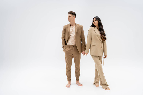 full length of barefoot interracial couple in beige pantsuits looking away on grey background - Foto, Imagem