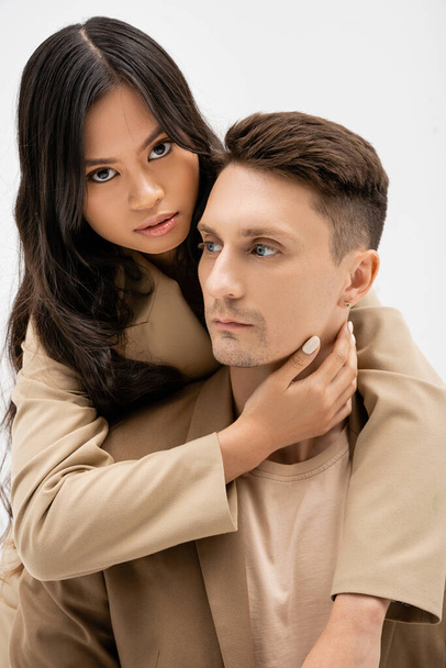 brunette asian woman looking at camera and hugging man in beige blazer isolated on grey - Photo, image
