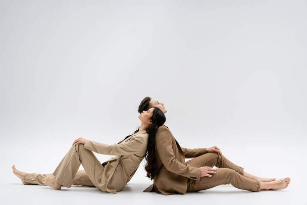 side view of multiethnic couple in beige suits sitting back to back on grey background - Foto, afbeelding