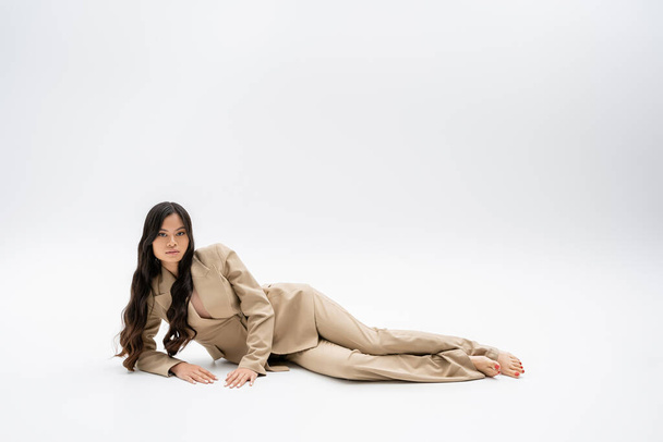 pełna długość boso azjatycki model w modny garnitur leżący i patrząc w aparat na szarym tle - Zdjęcie, obraz
