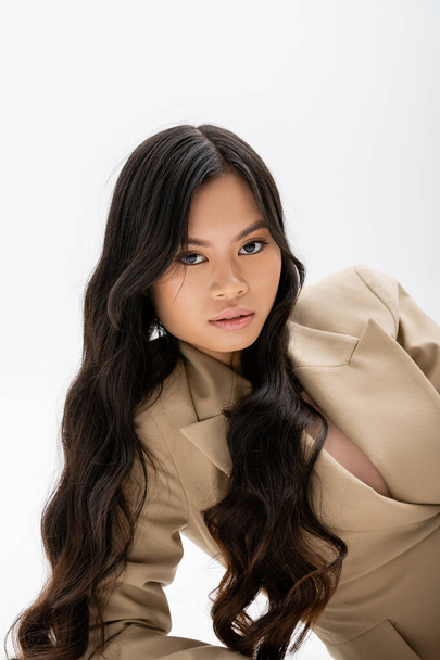 charming asian woman with long brunette hair looking at camera while posing in beige blazer on grey background - Foto, immagini