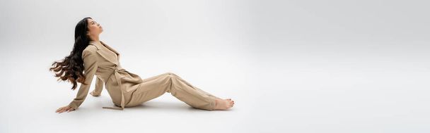 side view of barefoot asian woman with long brunette hair sitting in beige suit on grey background, banner - Foto, Imagem