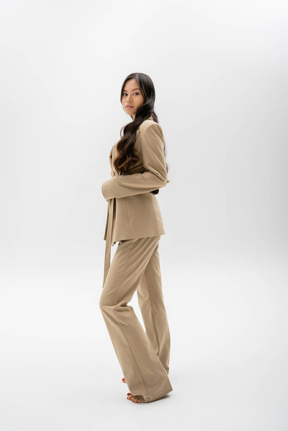 pleine longueur de pieds nus femme asiatique en veste beige et pantalon debout sur fond gris - Photo, image