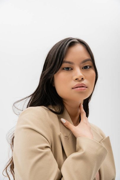 retrato de morena mujer asiática en blazer beige sosteniendo la mano cerca del cuello y mirando a la cámara aislada en gris - Foto, imagen