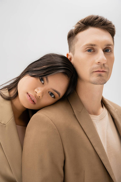 young asian woman leaning on stylish man in beige blazer and looking at camera isolated on grey - Photo, Image