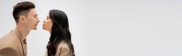 zijaanzicht van langharige Aziatische vrouw en trendy man poseren oog in oog met gesloten ogen geïsoleerd op grijs, banner - Foto, afbeelding