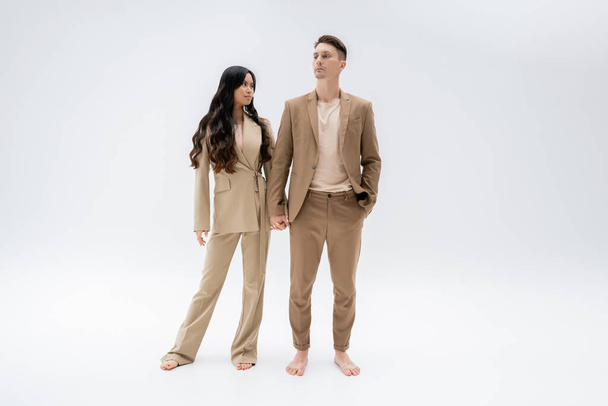 full length of barefoot multiethnic couple in trendy suits holding hands while standing on grey background - Foto, afbeelding