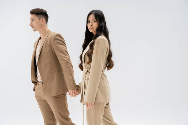 young asian woman with long hair looking at camera while holding hands with man in beige suit isolated on grey - Photo, Image