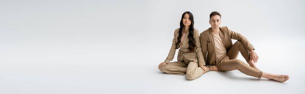 full length of barefoot interracial couple in beige suits sitting and looking at camera on grey background, banner - Foto, afbeelding