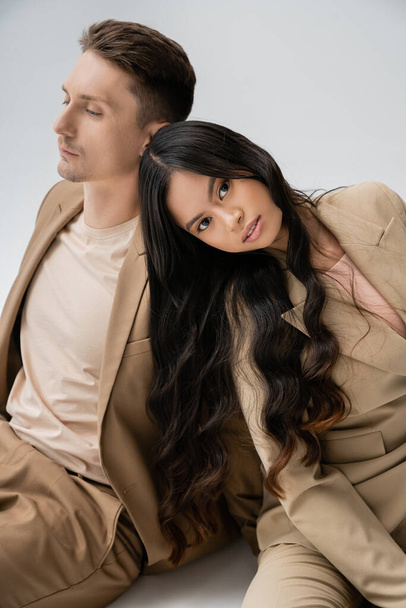 brunette asian woman with long hair looking at camera while sitting near man in beige suit on grey background - 写真・画像