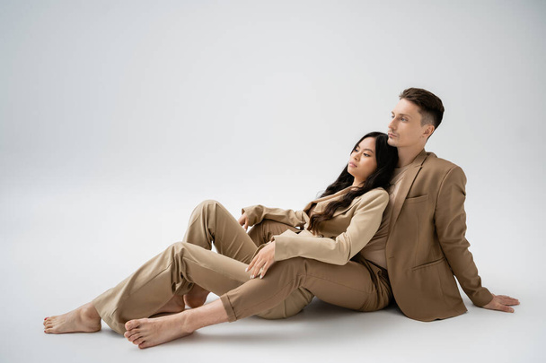 full length of barefoot interracial couple in beige pantsuits sitting on grey background - Fotó, kép