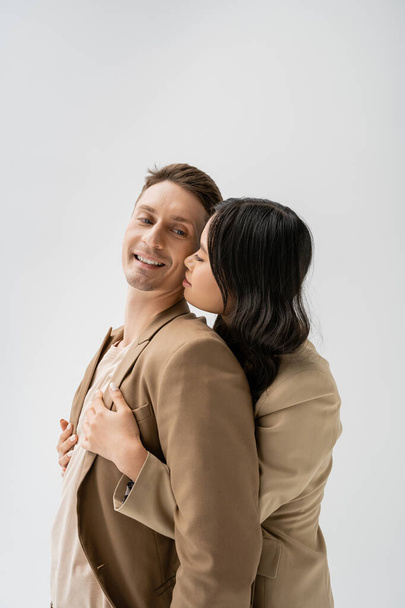 brunette asian woman embracing cheerful man wearing beige blazer isolated on grey - Foto, immagini