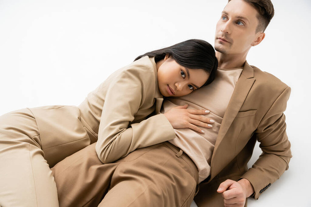 sensual asian woman looking at camera while hugging trendy man lying on white background - Photo, Image