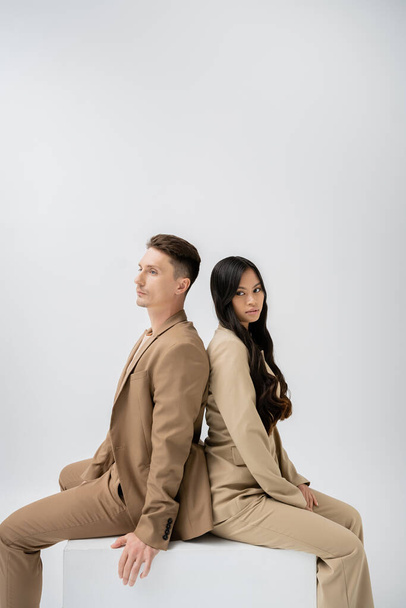 brunette asian woman with long hair looking at camera while sitting back to back with trendy man isolated on grey - Фото, изображение