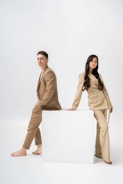full length of barefoot multiethnic couple in beige pantsuits looking at camera near white cube on grey background - Photo, Image