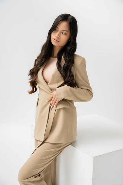 fashionable asian woman in beige blazer looking away near white cube on grey background - Zdjęcie, obraz