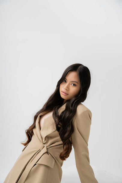 pretty asian woman with long brunette hair posing in stylish blazer and looking at camera isolated on grey - Photo, Image