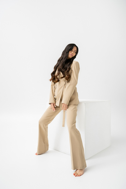 full length of barefoot asian woman in beige suit sitting on cube on white background - Φωτογραφία, εικόνα
