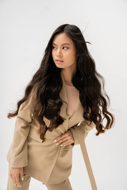 молодая азиатская женщина с длинными волосами брюнетки в бежевой куртке и глядя в сторону изолированы на сером - Фото, изображение