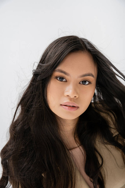 retrato de encantadora mujer asiática con pelo largo y saludable mirando a la cámara aislada en gris - Foto, imagen