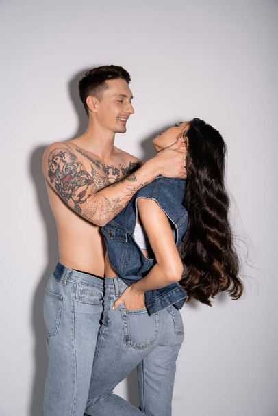 sonriente hombre con cuerpo tatuado sin camisa abrazando mujer asiática con pelo largo morena sobre fondo gris - Foto, Imagen