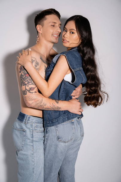 pleased man with shirtless tattooed body hugging brunette asian woman in denim outfit on grey background - Fotó, kép