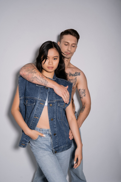 shirtless tattooed man embracing asian woman posing with hand in pocket of jeans on grey background - Photo, image