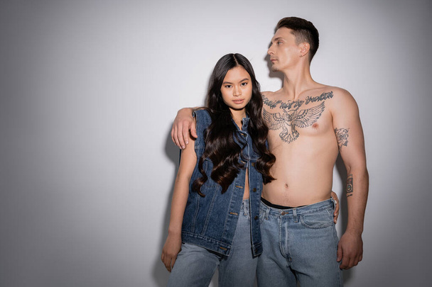 brunette asian woman in denim vest and jeans looking at camera near shirtless tattooed man on grey background - Fotó, kép