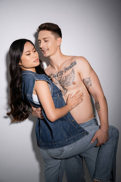 smiling tattooed man embracing sensual asian woman in denim outfit on grey background - Photo, Image