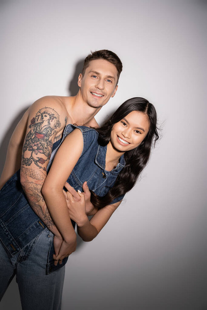 τατουάζ και shirtless άντρας αγκαλιάζει ευτυχισμένη Ασιάτισσα γυναίκα σε denim γιλέκο σε γκρι φόντο - Φωτογραφία, εικόνα