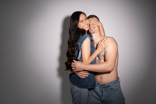 cheerful asian woman in denim clothes covering eyes of shirtless tattooed man on grey background - Foto, immagini
