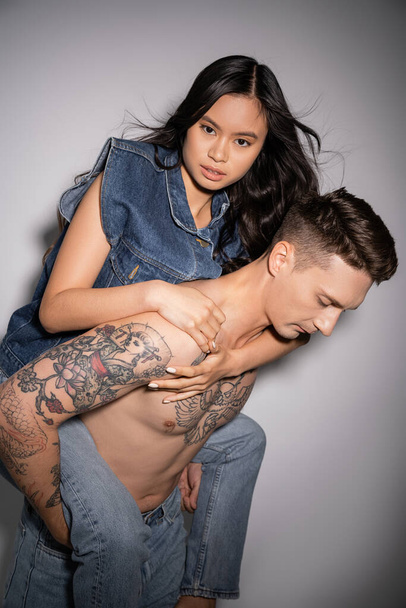 shirtless tattooed man piggybacking long haired asian woman in denim clothes on grey background - Photo, Image