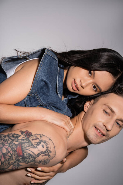 muscular and tattooed man piggybacking sensual asian woman looking at camera on grey background - Photo, Image