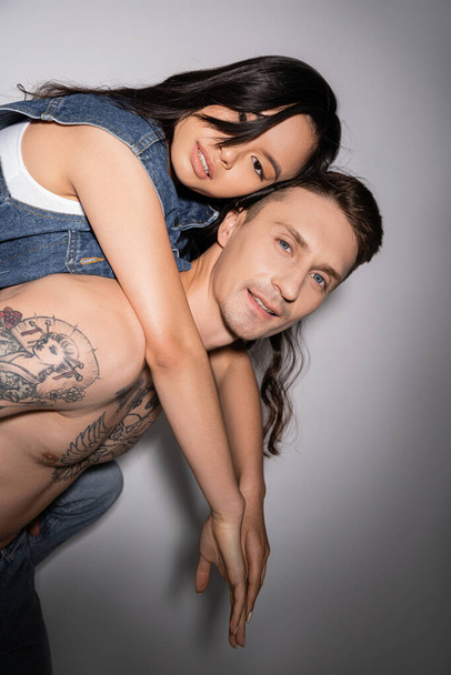 shirtless tattooed man and asian woman in denim vest looking at camera while having fun on grey background - Photo, Image