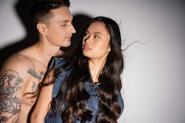 shirtless tattooed man and long haired asian woman in denim vest looking at each other on grey background - Fotoğraf, Görsel